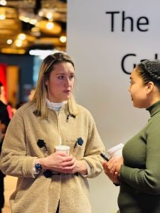 Two women stand in conversation at an event space named The Gallery. One woman, wearing a beige coat and holding a cup of coffee, listens attentively, while the other, dressed in a green sweater, also holds a coffee cup and gestures with a phone in hand.
